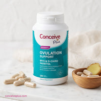 A bottle of Conceive Plus Women's Ovulation Support dietary supplement capsules, styled on a white marble surface with loose capsules and a small wooden bowl of ginger and capsules.
