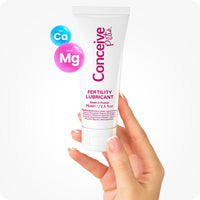Conceive Plus Fertility Lubricant Pack (ES) featuring 75ml multi-use tube, shown being held upright by a hand with manicured nails, alongside floating blue and pink ion symbols labeled Ca and Mg. White tube has pink branding text.