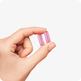 Conceive Plus Ovulation Bundle (ES) with a close-up of two pink Fertility Support capsules held gently between thumb and index finger, showing the capsule size and smooth texture for clear supplement visualization.