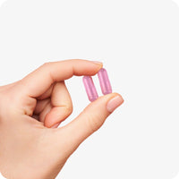 Conceive Plus Ovulation Bundle (ES) with a close-up of two pink Fertility Support capsules held gently between thumb and index finger, showing the capsule size and smooth texture for clear supplement visualization.